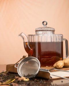 Elegant glass teapot filled with tea, surrounded by ginger and tea leaves on a rustic table.