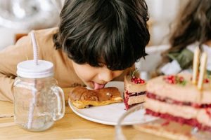 Young boy playfully licking a chocolate eclair at a birthday party table.